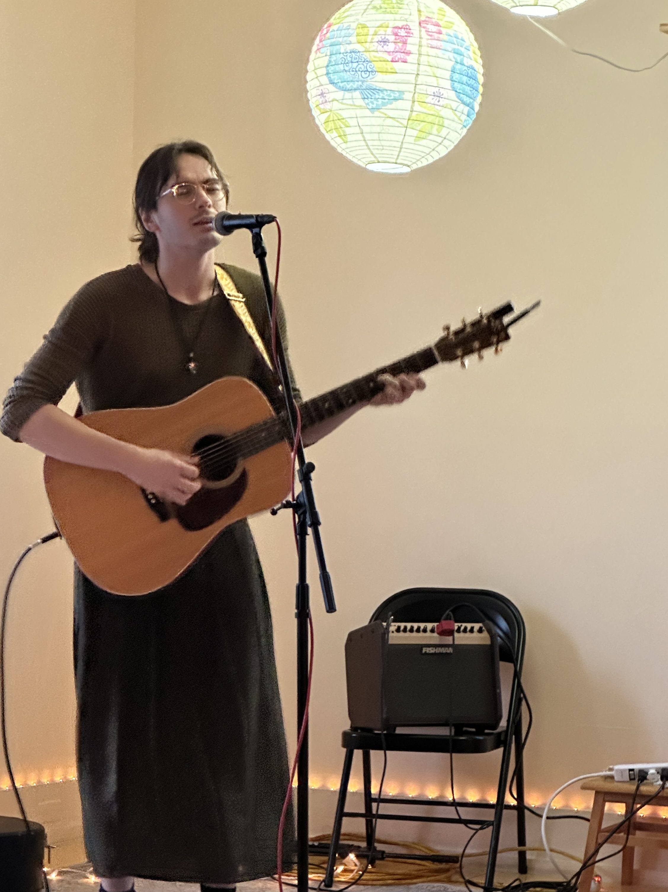 A musician performing live, playing an acoustic guitar and singing into a microphone, with stylish ambient lighting and decorative globes in the background.